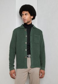 Chaqueta de botón verde texturizada con cuello, con un bolsillo en el pecho, llevada sobre un suéter negro de cuello alto y pantalones beige.