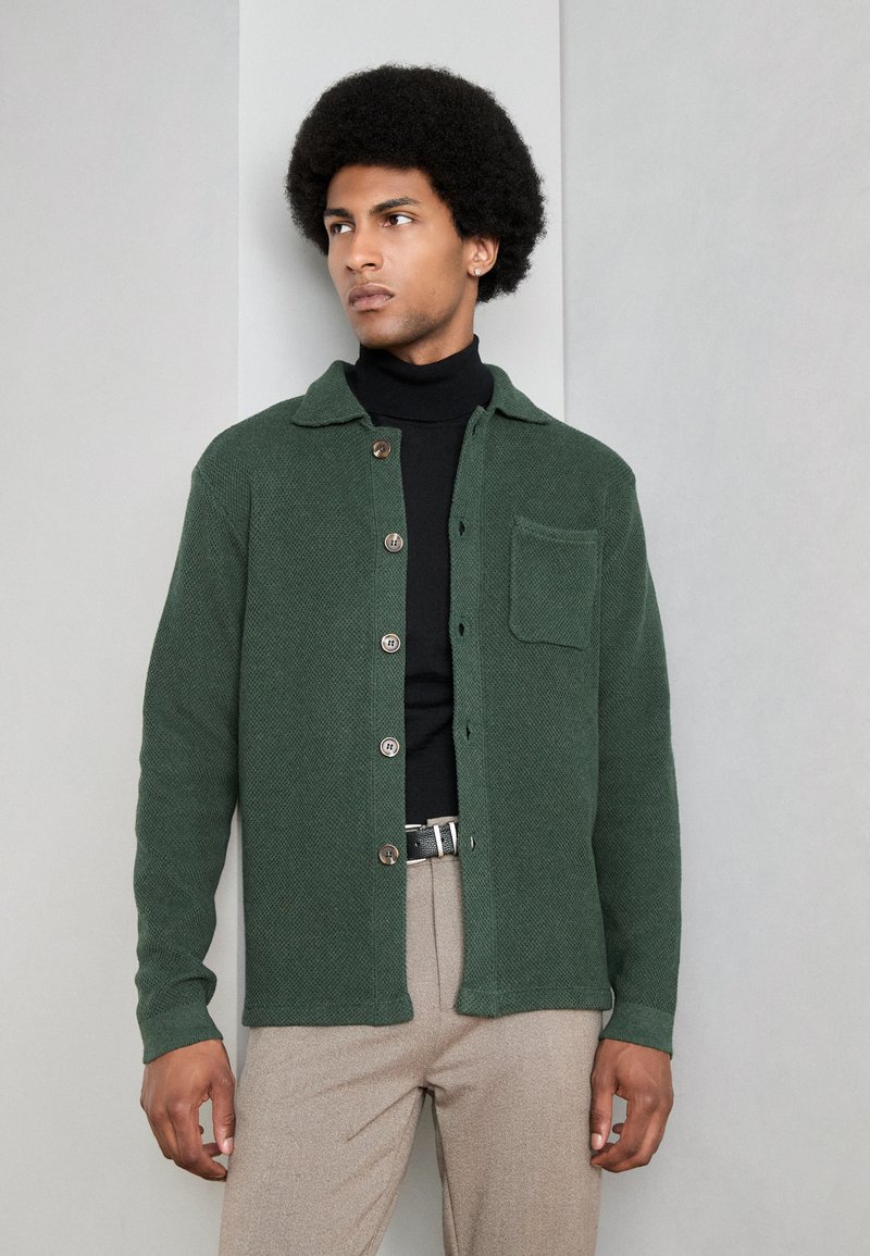 Chaqueta de botón verde texturizada con cuello, con un bolsillo en el pecho, llevada sobre un suéter negro de cuello alto y pantalones beige.