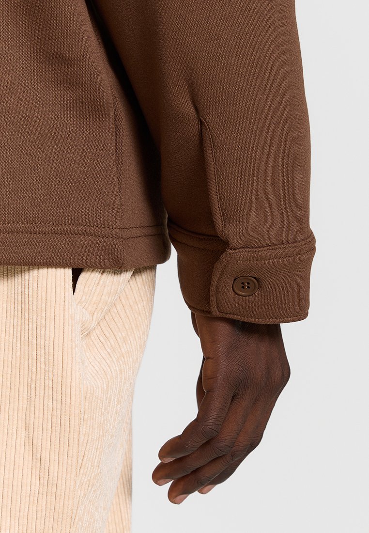 Gros plan d'une main à la peau foncée reposant à côté d'un pantalon en velours côtelé beige et d'une chemise à manches longues marron avec un poignet boutonné.