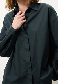 Camicia button-up oversize verde scuro con colletto pieghevole, maniche lunghe e una texture liscia. Bottoni distintivi e vestibilità rilassata.