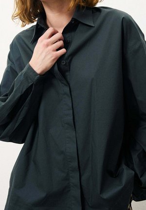 Camisa abotonada de gran tamaño en verde oscuro, con cuello doblado, mangas largas y una textura suave. Botones notables y ajuste relajado.