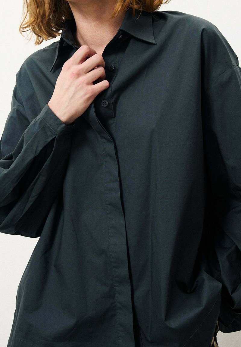Camicia button-up oversize verde scuro con colletto pieghevole, maniche lunghe e una texture liscia. Bottoni distintivi e vestibilità rilassata.