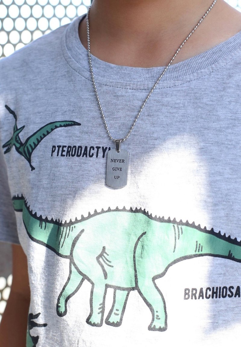 Grijze t-shirt met dino-afbeeldingen in groen en zwart; zilveren ketting met rechthoekige hanger met de inscriptie "GEEN OPGEVEN."