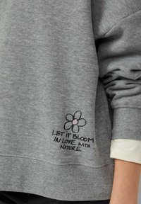 Felpa grigia con una texture morbida. Presenta un fiore stampato con un centro rosa e il testo "LASCIA CHE FIORISCA NELL'AMORE PER LA NATURA" sotto.