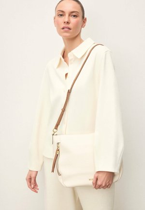 Femeie purtând o jachetă crem cu nasturi și pantaloni, ținând o geantă mare de umăr de culoare alb-crem cu o curea reglabilă maro și fermoar auriu.