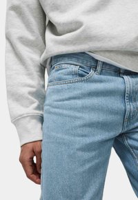 Jeans de mezclilla azul claro con corte Slim, que presentan un ligero efecto de desgaste, costuras naranjas y un bolsillo delantero visible. Acompañados de una sudadera gris.