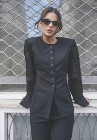 Blazer nero su misura con maniche lunghe in pizzo, colletto rotondo e sei bottoni davanti, abbinato a pantaloni neri.