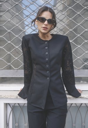 Blazer nero su misura con maniche lunghe in pizzo, colletto rotondo e sei bottoni davanti, abbinato a pantaloni neri.