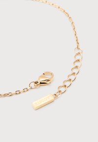 Χρυσή αλυσίδα με μικρούς ωοειδείς κρίκους, κούμπωμα καβουράκι, επέκταση αλυσίδας και ορθογώνια ετικέτα με χαραγμένο το "LACOSTE" σε γκρι φόντο.