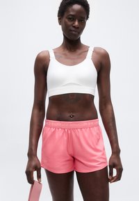 Αθλητική γυναίκα με λευκό αθλητικό σουτιέν και ροζ σορτς Under Armour κρατώντας μια ροζ ταινία αντίστασης μπροστά σε απλό φόντο.