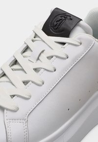 Λευκό δερμάτινο αθλητικό παπούτσι με λευκά κορδόνια και μαύρη ετικέτα στη γλώσσα με ανάγλυφο λογότυπο.