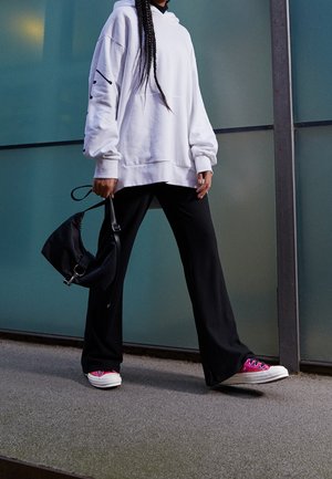 Person wearing oversized white hoodie, black wide-leg pants, pink sneakers, holding a black bag, standing against glass wall outdoors.