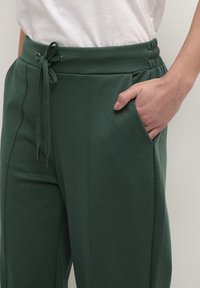 Pantalons de jogging verts en tissu doux, dotés d'une taille à cordon, de poches latérales et d'une coupe décontractée. La surface est lisse, sans motifs visibles.
