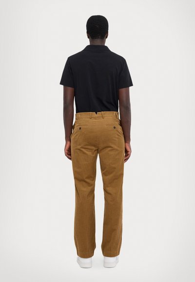 Polo noir, pantalon en velours côtelé brun avec une texture lisse, coupe droite et deux poches arrière, porté avec des baskets blanches.