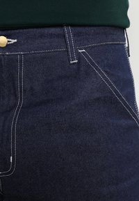 Dark blue denim jeans with contrasting white stitching, five-pocket design, and a metal button at the waist. Smooth texture.