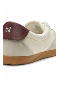 Baskets crème et beige avec semelle en gomme, languette du talon bordeaux cousue avec la marque "HUGO", vues sous un angle arrière latéral.