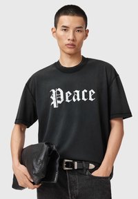 Fekete rövid ujjú póló "Peace" felirattal fehér gótikus betűkkel, fekete bőrtáskával és övvel párosítva, semleges háttér előtt.