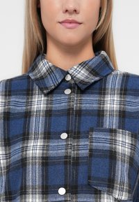Blå og svart rutete flanellskjorte med krage, metallknapper og brystlomme. Myk tekstur, rutete mønster, skreddersydd design.
