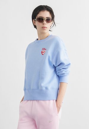 Isik, kellel on päikeseprillid, helesinine dressipluus punase südame logoga ja heleroosad Nike dressipüksid, käed taskutes, vaadates küljele.