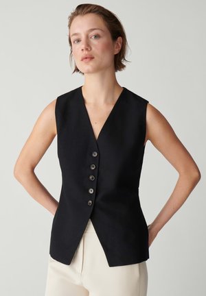 Junge Frau mit kurzen Haaren trägt eine ärmellose schwarze, geknöpfte Weste und hellfarbene Hose, steht vor einfachem Hintergrund mit Händen in der Hüfte.