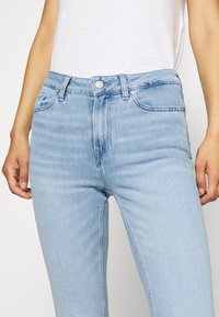 Jeans de mezclilla azul claro con cintura alta, diseño de cinco bolsillos, costuras en contraste y cierre de botón frontal. Textura suave y ajuste slim.