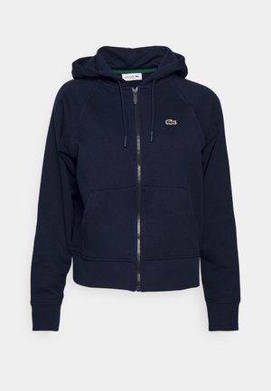 Sudadera con cremallera de color azul marino en tejido suave, con capucha, bolsillos frontales y un pequeño logo de Lacoste en el lado izquierdo del pecho. Puños y dobladillo de canalé.