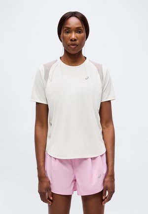 Femme portant un t-shirt de sport blanc à manches courtes et un short de course rose clair, debout devant un fond blanc uni.
