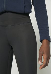 Mallas negras de cintura alta, hechas de un material elástico con costuras planas. Se ve una mano dentro de la manga de un sudadera color marino.