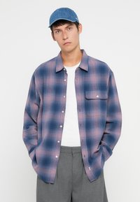Plaid button-up shirt in shades of blue and pink, featuring a chest pocket and relaxed fit. Paired with a plain white t-shirt and gray trousers.