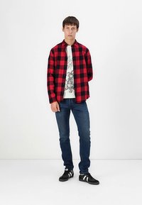 Κόκκινο και μαύρο πουκάμισο με καρό σχέδιο πάνω από λευκή γραφιστική μπλούζα, συνδυασμένο με σκούρο μπλε τζιν και μαύρα αθλητικά παπούτσια με λευκές λεπτομέρειες.