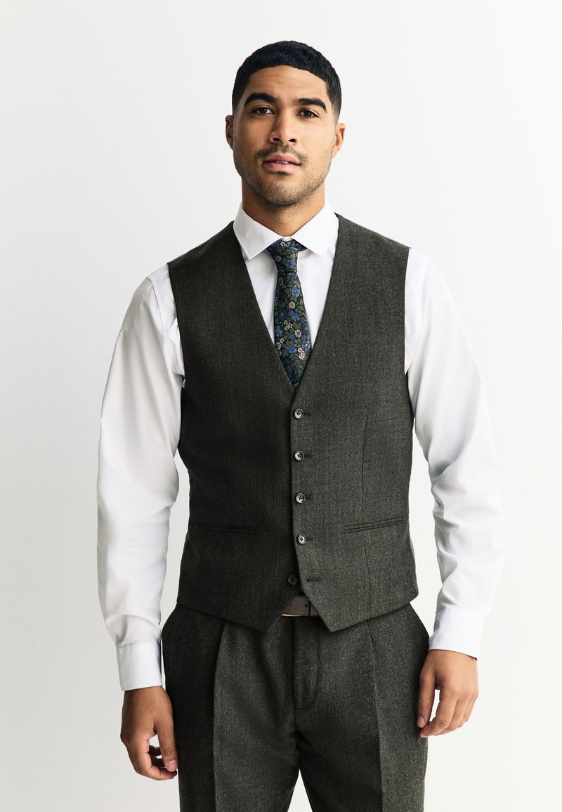 Homme portant un gilet gris foncé boutonné, un pantalon assorti, une chemise blanche et une cravate à motif floral, debout devant un fond clair uni.