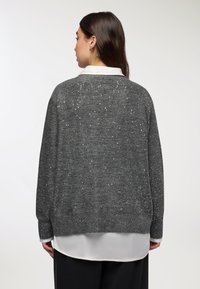 Szary, błyszczący sweter o luźnym kroju, z teksturowaną tkaniną i srebrnymi drobinkami. Stylizowany na białej bluzce z kołnierzykiem i czarnych spodniach.