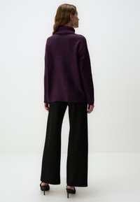 Losse turtleneck trui in diep paars, gebreide textuur, gecombineerd met zwarte wijde broek en zwarte hoge hakken.