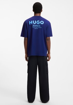 Mand iført en blå T-shirt med stor "HUGO" tekst og romertal på bagsiden, kombineret med løse sorte cargobukser, stående med ryggen til.