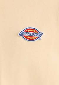 Bežinė trumpa rankovė marškinė su raudonu ir mėlynu audiniu išsiuvinėtu "Dickies" logotipu, esančiu kairėje krūtinės pusėje. Minkšta, lygi audinio tekstūra.