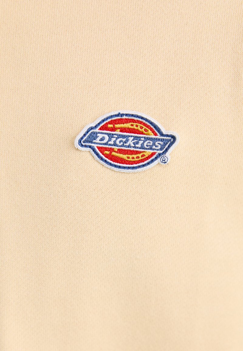T-shirt beige à manches courtes avec un logo Dickies tissé en rouge et bleu, placé sur la partie gauche de la poitrine. Texture de tissu douce et lisse.