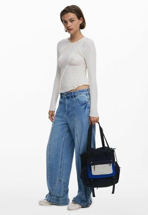White long-sleeve top with textured pattern, paired with loose blue denim jeans. Model holds a multicolored tote bag with various pockets.