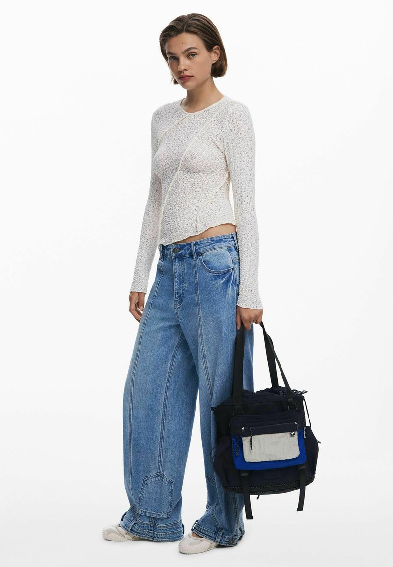 Haut à manches longues blanc avec un motif texturé, associé à un jean en denim bleu ample. Le modèle tient un tote bag multicolore avec plusieurs poches.