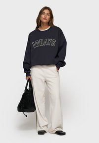 Felpa navy con logo "10DAYS", pantaloni crema bianchi, borsa nera e scarpe nere. Vestibilità oversize, texture morbida, colori a contrasto.