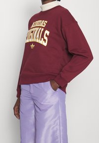 Vinröd sweatshirt med "Adidas Originals"-logotyp i gul text, löst snitt, rund hals, ribbade manschetter, matchad med glänsande lavendelfärgade byxor.