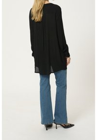 Chemise noire à manches longues en tissu texturé, avec un pli au dos, portée avec un jean flare bleu clair et des escarpins noirs à bouts pointus.