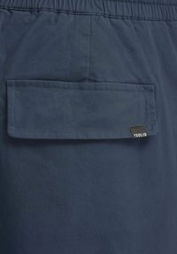 Navy shorts made of lightweight fabric, featuring a patch pocket with a flap, elastic waistband, and a small black branded label.