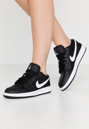 Nogi w czarno-białych butach Nike Air Jordan stoją na gładkiej białej podłodze na jednolitym tle.