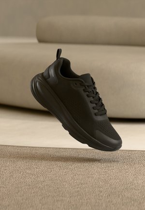 Černá sportovní teniska s tkaničkami vznášející se nad béžovým kobercem v minimalistické místnosti s béžovou pohovkou v pozadí.