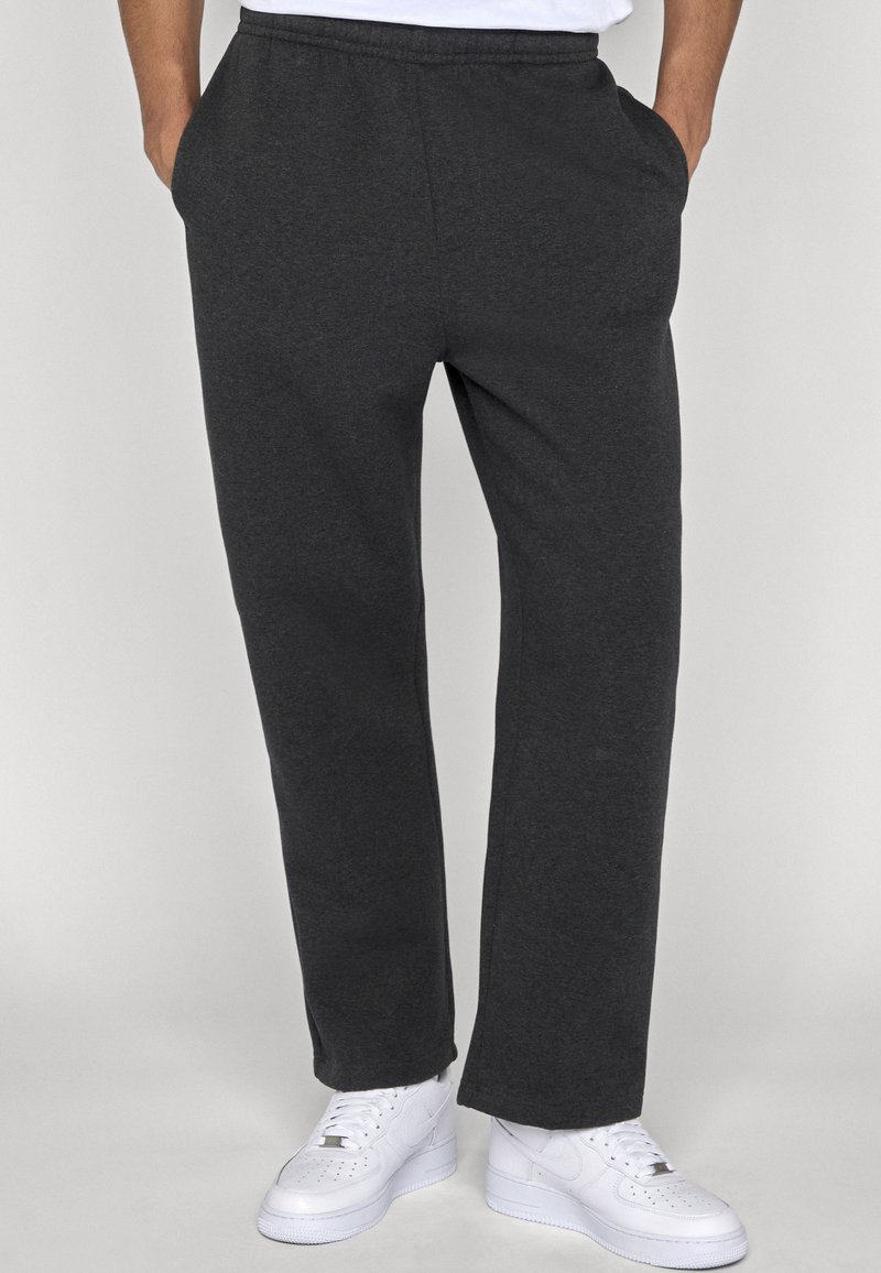 Personne portant un pantalon de survêtement gris foncé avec des poches latérales et des baskets blanches, debout les mains dans les poches devant un fond uni.