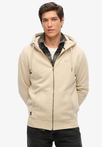 Sudadera beige de cremallera con capucha ajustable, bolsillos frontales y puños acanalados, superpuesta a una camisa de cuadros. El tejido parece suave y liso.