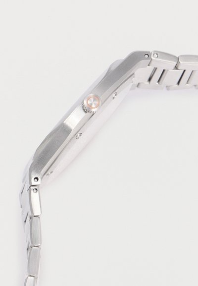 Ασημένιο μεταλλικό ρολόι χειρός, πλευρική όψη με έμφαση στην κορόνα και τον ενωμένο βραχίονα, σε λευκό φόντο.