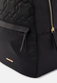 Mochila acolchada negra con textura suave, cremallera tono dorado y un bolsillo delantero con un diseño elegante y acentos delgados.