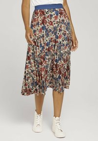 Jupe plissée florale avec une taille bleu, présentant des motifs rouges, bleus et noirs sur fond beige. Assortie à des bottines blanches.