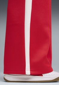 Red track pants with white side stripes, made from textured fabric. A white and gray sneaker with brown soles is partially visible underneath.